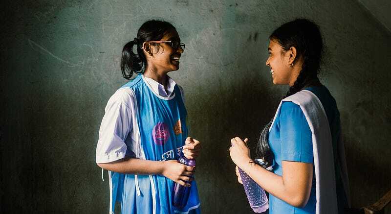 Two girls chat and smile in a hallway. Both hold water bottles and are wearing Splash vests.